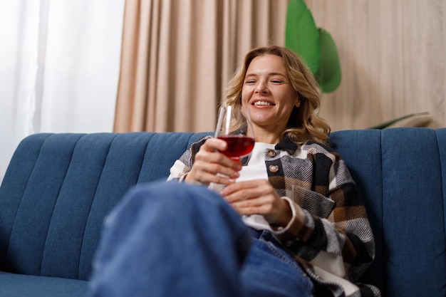 Happy middle aged caucasian woman holding glass of red wine on sofa