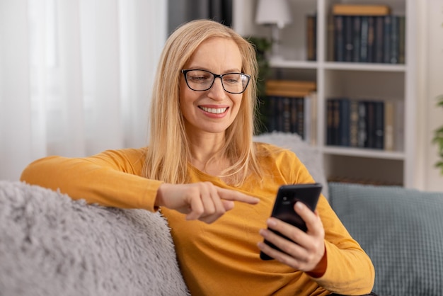 Happy middle aged lady resting on cozy grey sofa and surfing internet on modern cell phone Caucasian woman enjoying free time at home with smartphone in hands