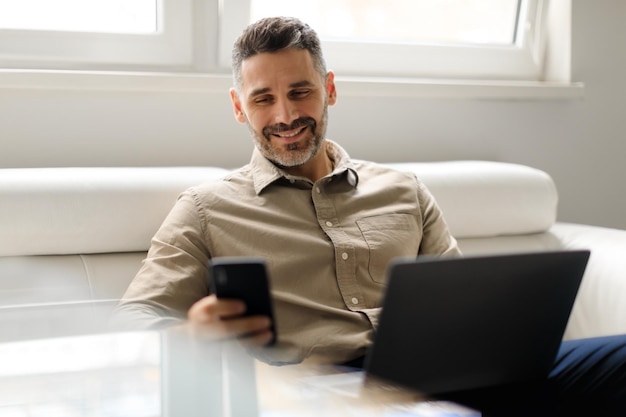 Happy middle aged male ceo holding smartphone and using laptop sitting in office interior on couch free space