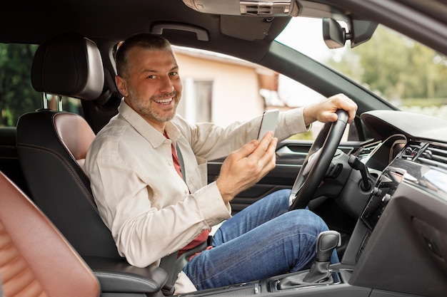 Happy middle aged man driver using smartphone while sitting in car at driver seat using navigation