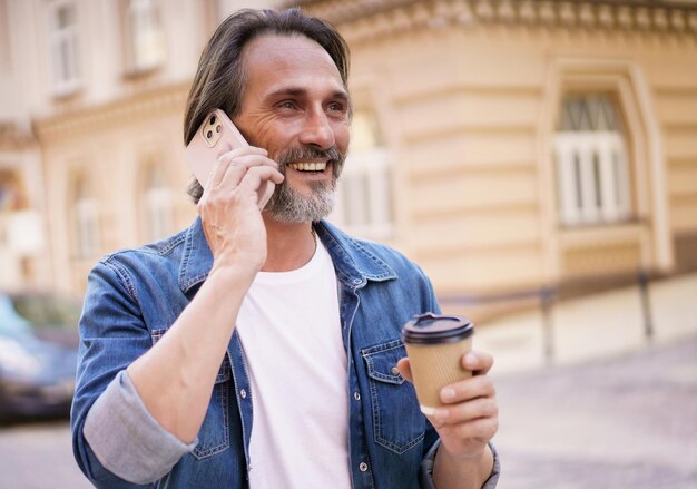 Happy middle aged man with grey bearded talking on the phone holding coffee in disposable paper cup standing outdoors in old city background wearing jeans shirt Freelancer traveling man