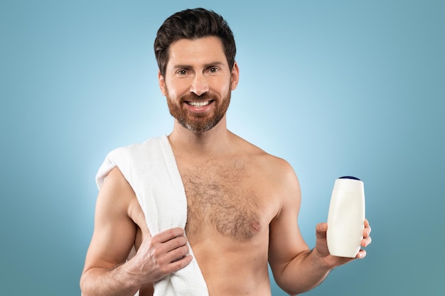 Happy middle aged man with towel on shoulder holding bottle with moisturising body lotion or shampoo mockup