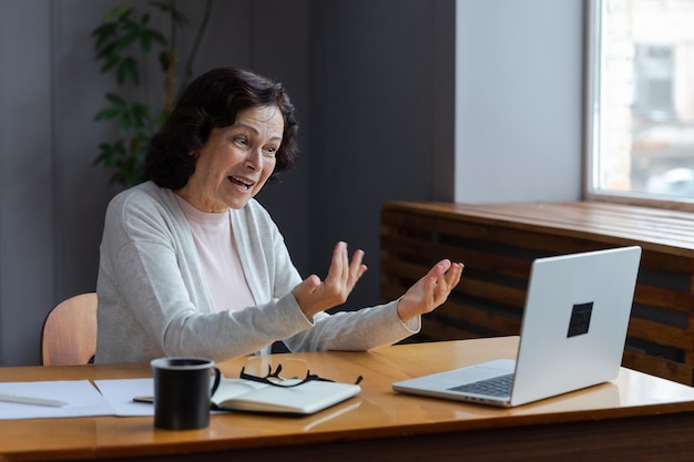 Happy middle aged senior woman sit with laptop talk on video call with friends family laughing matur