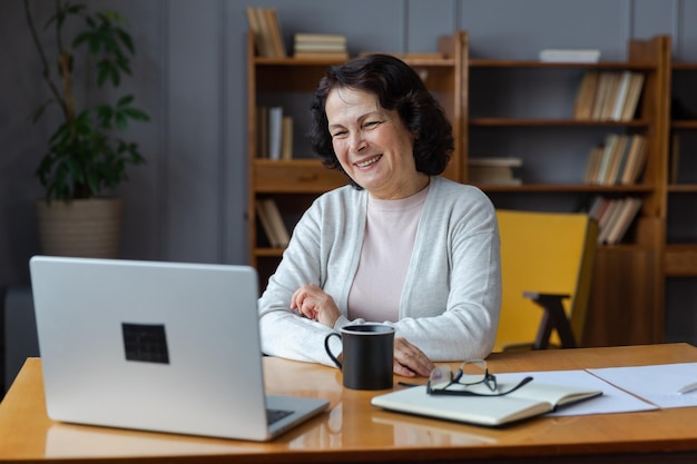 Happy middle aged senior woman sit with laptop talk on video call with friends family laughing matur