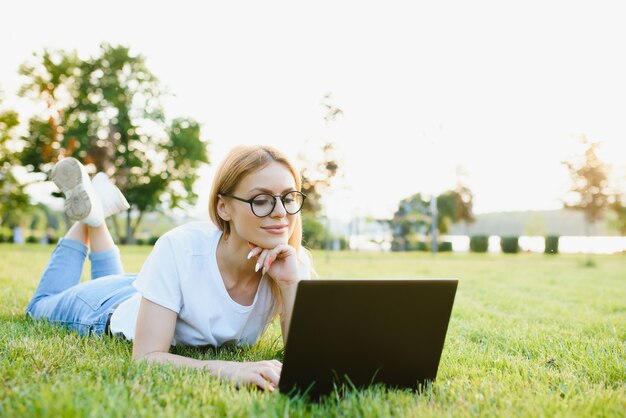 Happy middle aged woman lying on green grass using laptop computer in the park