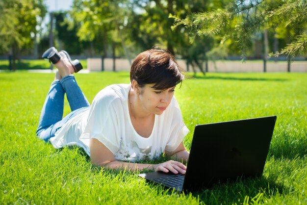 Happy middle aged woman lying on green grass using laptop computer in the park.