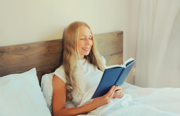 Счастливая женщина среднего возраста читает книгу, лежа в постели дома.