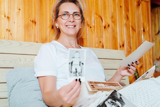 Happy middle aged woman showing old photo of childhood while relaxing on bed in bedroom and looking at camera. Adult woman and vintage pictures, retro photo from family album. Nostalgia and memories.