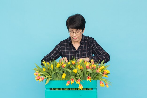 Happy middle-aged woman with glasses holding box of tulips in blue background