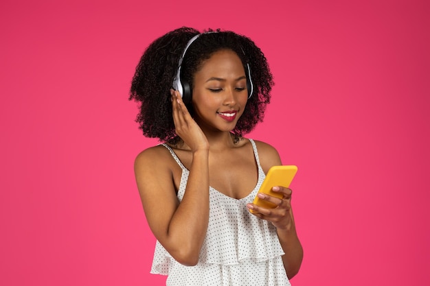 Happy millennial african american curly lady in wireless headphones listening to music typing on