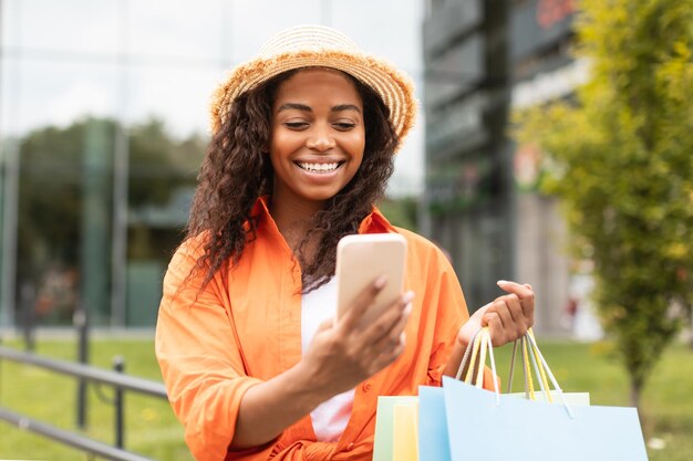 Happy millennial african american lady in casual and hat with\
many bags chatting on smartphone in city street