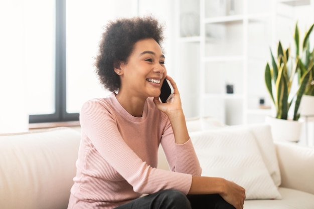 Happy millennial african american woman calling by smartphone talking sitting on sofa