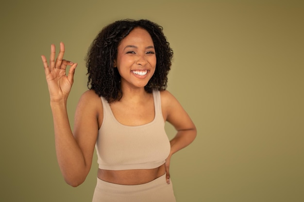 Happy millennial african american woman in sportswear show ok sign isolated on beige studio