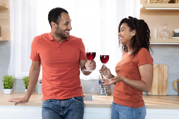 Happy millennial black lady and guy with glasses of red wine cheers toast talking and enjoy free time