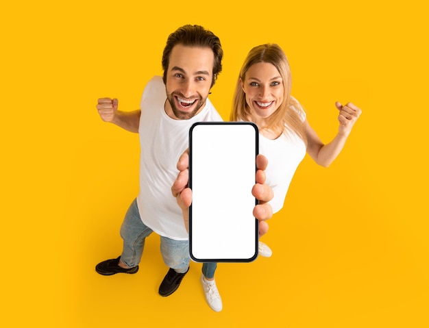 Happy millennial caucasian couple in white tshirts enjoy winning online show smartphone with blank screen