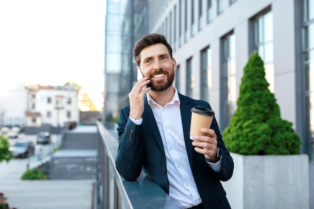 Happy millennial european male with beard in suit call by smartphone drinks from cup takeaway on balcony