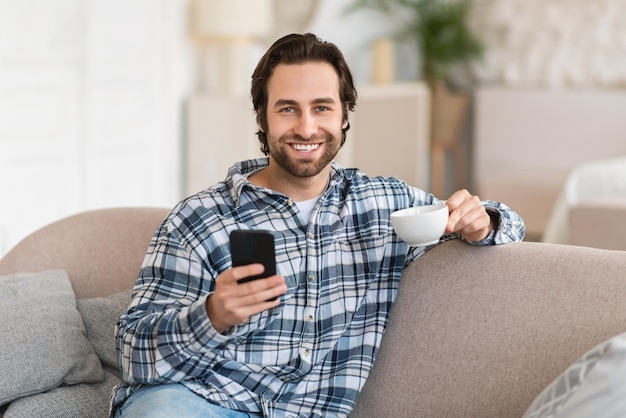 Happy millennial european man in domestic clothes with stubble chatting on smartphone watch video