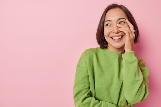 Happy millennial girl with dark hair covers face with hand\
smiles joyfully looks aside feels carefree dressed in casual green\
jumper isolated over pink background empty space for your\
promotion