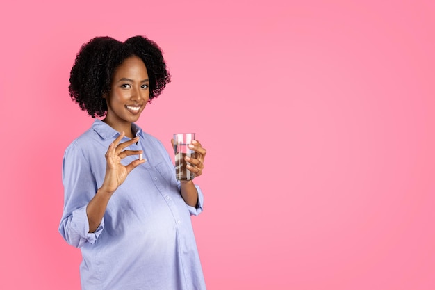 큰 배를 가진 행복한 밀레니엄 임신 흑인 여성은 알약 캡슐과 물 한 잔을 보여줍니다