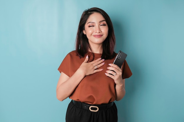 Happy mindful thankful young woman holding phone and hand on\
chest smiling isolated on blue background feeling no stress\
gratitude mental health balance peace of mind concept