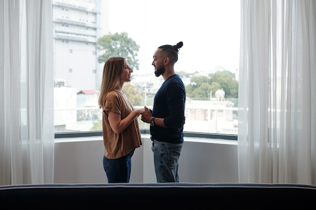 Happy mixed-race couple in love standing on balcony, holding hands and looking at each other