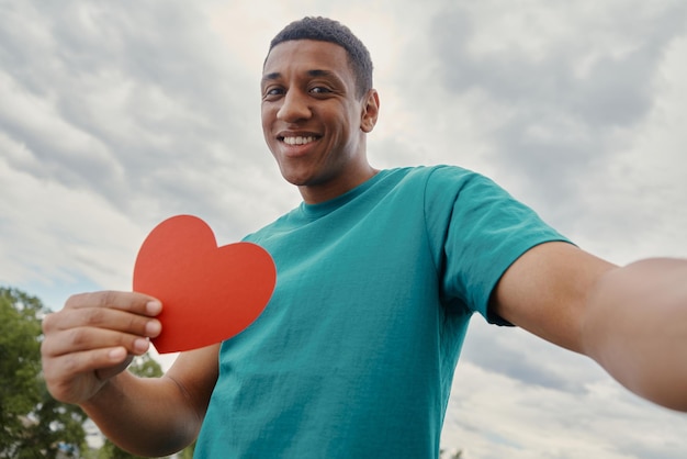 Uomo di razza mista felice che fa selfie e tiene a forma di cuore di carta con il cielo sullo sfondo