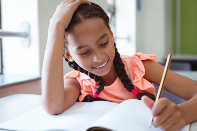 Happy mixed race schoolgirl in classroom sitting at desk smiling and writing in book