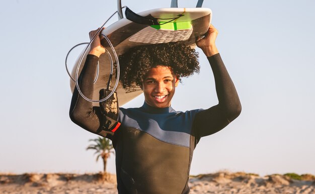 Счастливый мальчик-серфер смешанной расы балансирует доску для серфинга на голове. Смотрит прямо в камеру и улыбается.