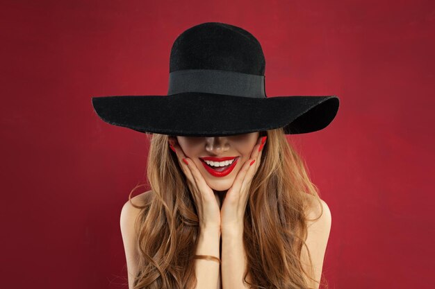写真 赤い背景で赤い唇のメイクアップとマニキュアをしている幸せなモデル女性 黒い帽子のモデル 肖像画