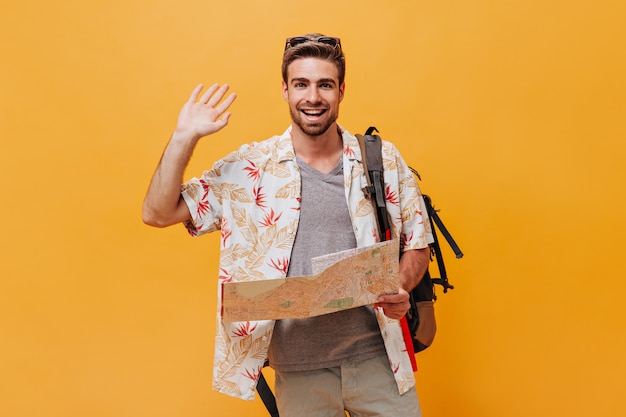 Happy modern guy with cool hairstyle and beard in grey t-shirt and white printed summer shirt holding map and greeting on isolated wall