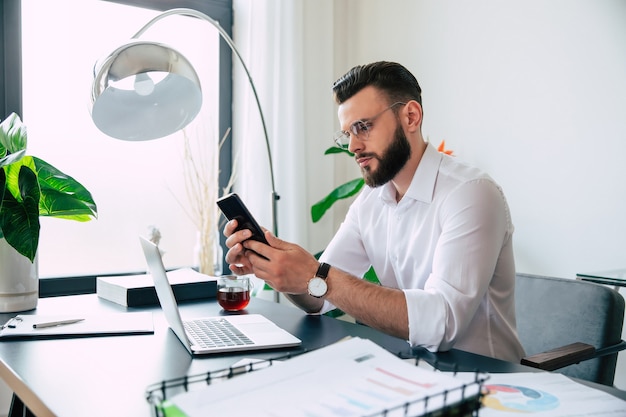 행복 한 현대 잘 생긴 세련 된 수염 된 사업가 안경에 흰색 셔츠는 사무실 직장의 책상에 노트북 및 smert 전화에서 작동합니다.
