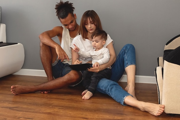 A happy modern multiracial family with a little boy sits against a gray wall in a room looks into a digital tablet