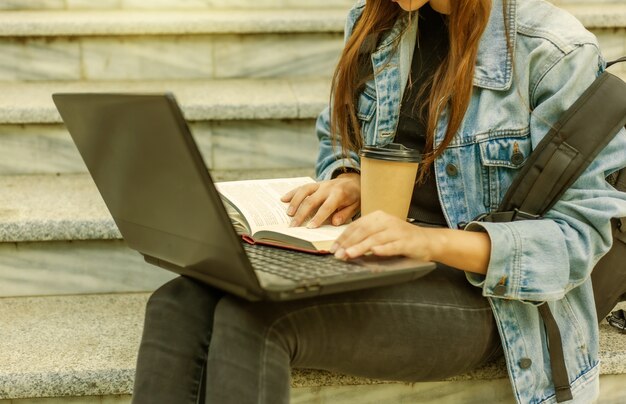 Felice studentessa moderna in giacca di jeans e zaino seduto sulle scale e usa il computer portatile con il libro all'aperto. insegnamento a distanza. concetto di gioventù moderna.