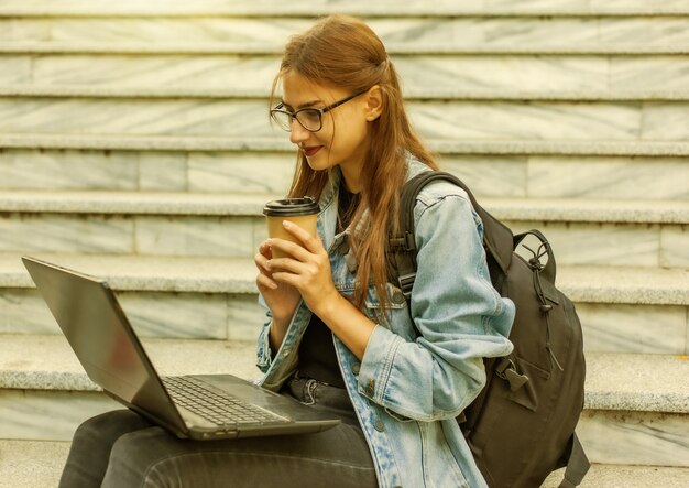 屋外のラップトップで階段に座っているデニムジャケットとバックパックで幸せな現代の女性の学生。コーヒーを飲む。ビデオを見る。通信教育。現代の若者のコンセプト。