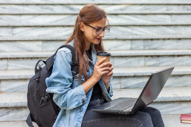 Studentessa moderna felice in rivestimento e zaino del denim che si siedono sulle scale con il computer portatile all'aperto. bere caffè. guardare video insegnamento a distanza. concetto di gioventù moderna.