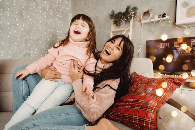 Happy mom and daughter jumping on the sofa together holding hands, mother playing with cute little girl at home, young mom and baby love to spend time together.