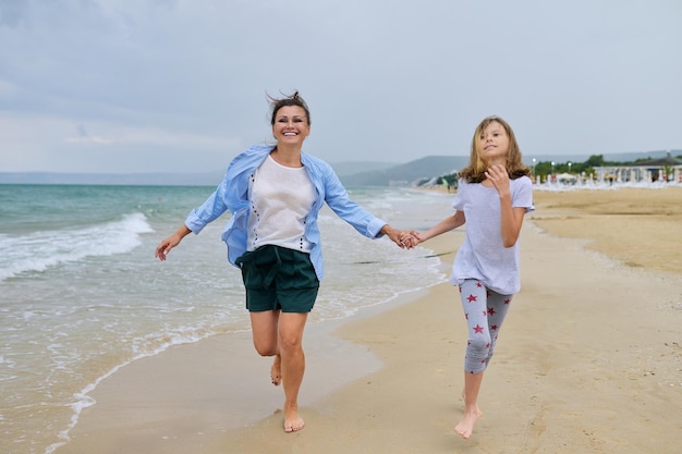 Happy mom and daughter running on the sea beach holding hands