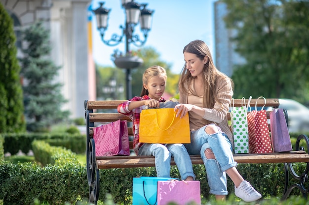 happy mom and daughter sitting on park bench, taking out item from shopping bag