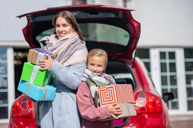 Happy mom and daughter standing back to back with gift boxes at car trunk