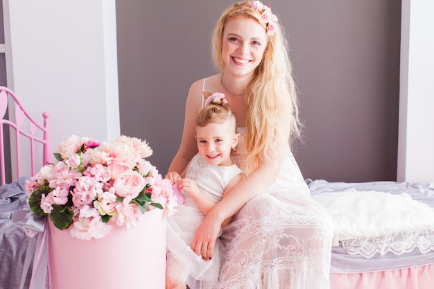 Happy mom and daughter in white dresses sitting on bed next to a bouquet of flowers