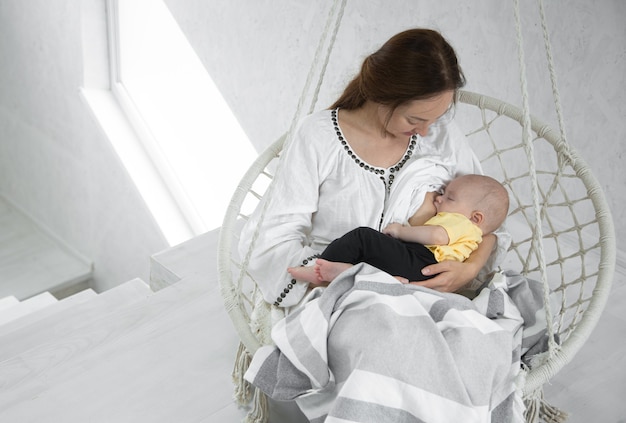 La mamma felice alimenta un bambino su un'amaca bianca con una coperta di una stanza bianca. concetto di maternità felice.