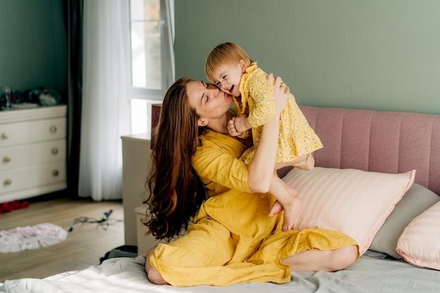 Happy mom hugging and kissing toddler daughter in the bedroom in the morning