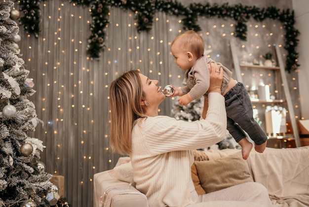 Happy mom is sitting on the couch and playing with a baby boy in a New Year's decor