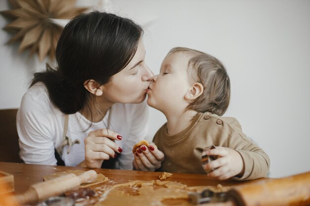 一緒にクリスマスのジンジャーブレッド クッキーを作る愛らしい幼児の娘にキスをする幸せなお母さん