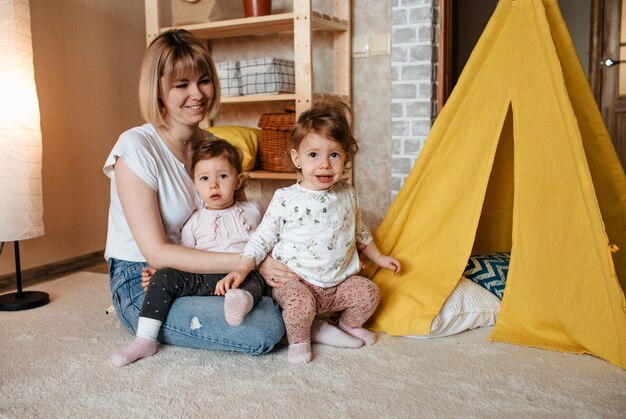 2人の双子の女の子と床で遊んで幸せなお母さん。黄色い小屋