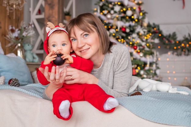 Mamma felice che gioca con il suo bambino in costume rosso da renna santa sullo sfondo dell'albero di natale