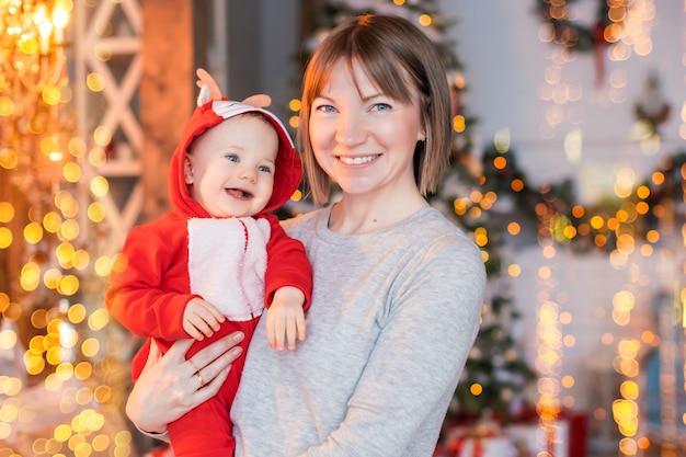 크리스마스 트리 배경에 빨간 산타 순록 의상을 입고 아기와 함께 노는 행복한 엄마