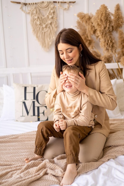 La mamma felice gioca a peekaboo con un bambino piccolo che chiude gli occhi su un letto nei toni naturali del marrone beige che si diverte insieme