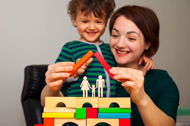 Happy mom and son are playing and building a house for the family. They put a roof. A woman shows and teaches the boy how important it is to quarantine at home.