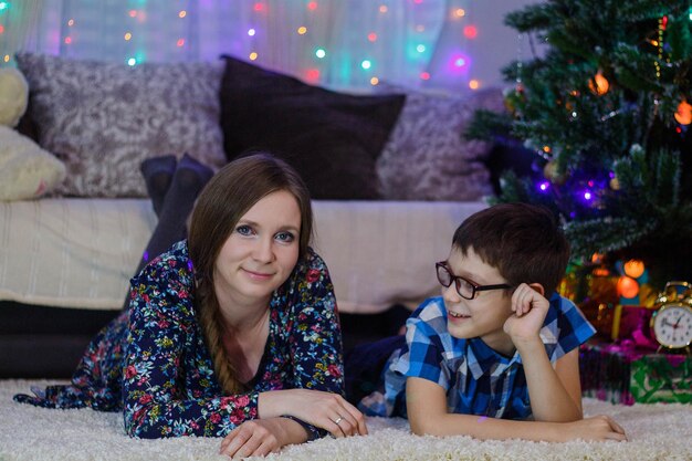 집에서 행복한 엄마와 아들이 하얀 카펫에 누워 새해 분위기 크리스마스 트리와 선물